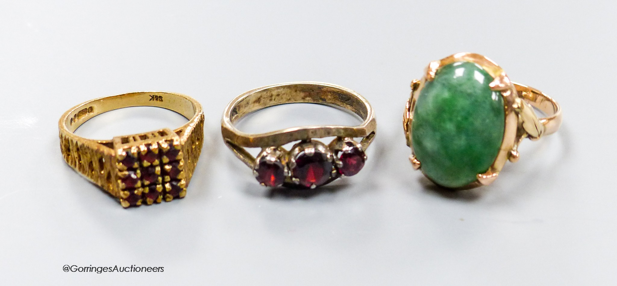 A 9ct gold and garnet cluster ring, a similar white metal ring and a yellow metal and jadeite cabochon ring, gross weight 12.1 grams.